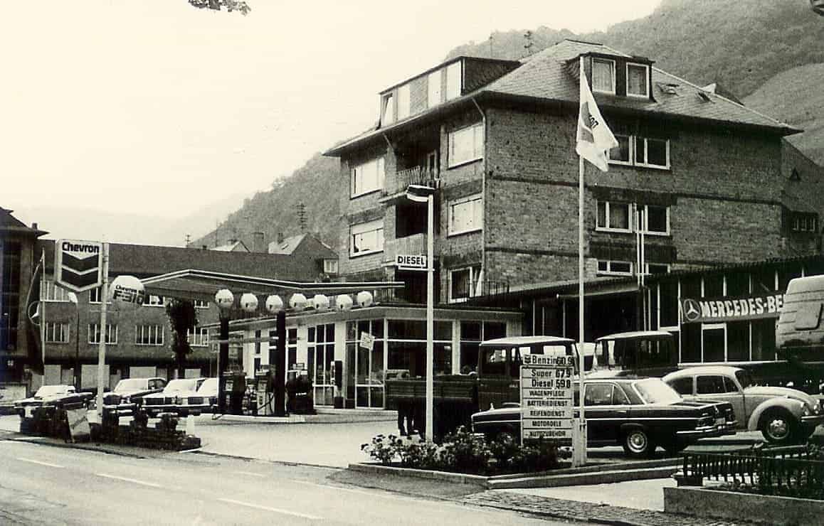 Neu errichteter Standort in 1967 mit Tankstelle mit Chevron-Schild, mehreren Zapfsäulen und mehreren geparkten Autos. Im Hintergrund steht ein größeres Gebäude mit dem Schriftzug ‚Mercedes-Benz‘. Ein Fahnenmast mit Flagge sowie ein Preisschild mit folgenden Angaben sind sichtbar: ‚Super 0,57 – Diesel 0,51 – Waschdienst – Reifenservice – Abschleppdienst – Motorölwechsel – Autovermietung‘. Im Hintergrund sind weitere Gebäude und Berge zu sehen.