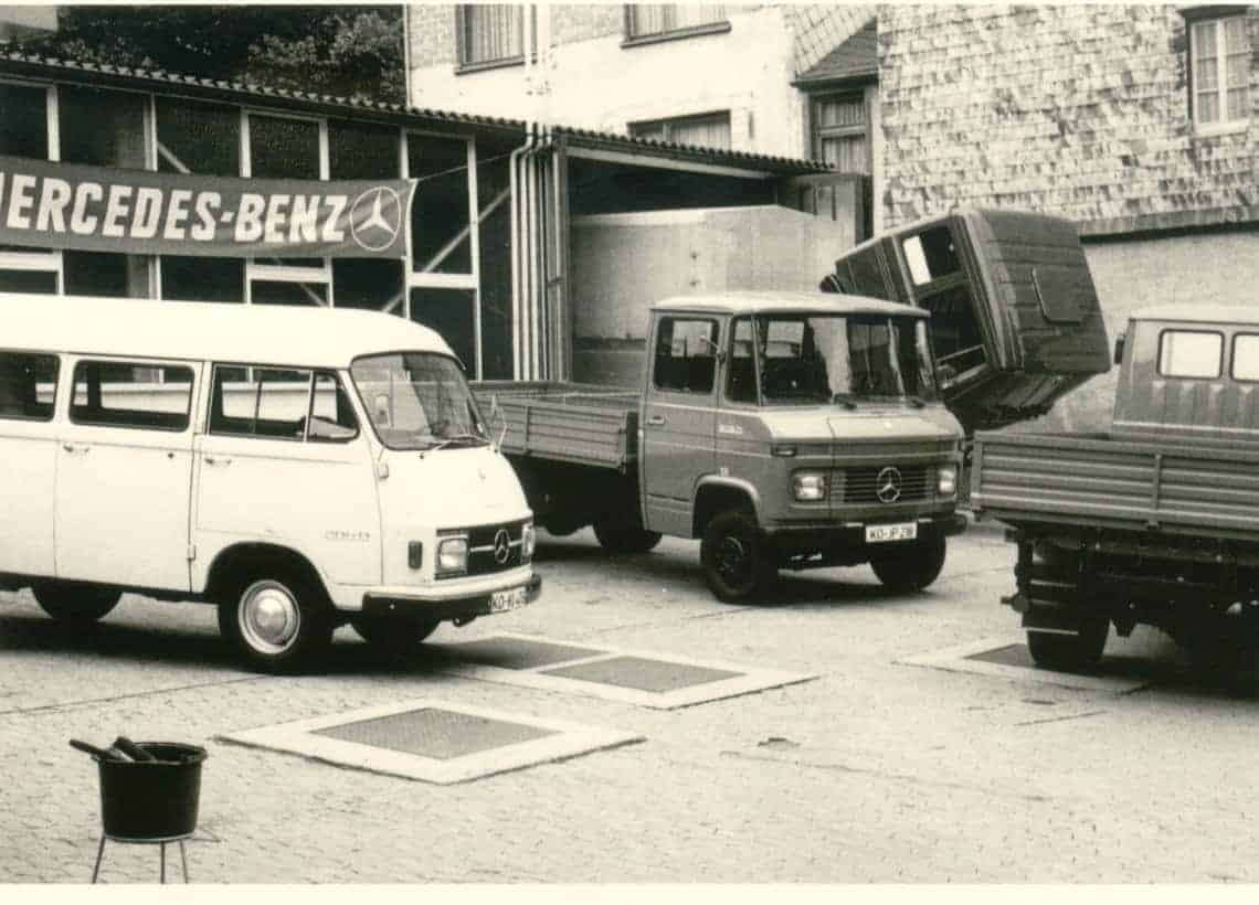 Schwarz-weiß-Fotografie mit mehreren Mercedes-Benz-Fahrzeugen vor einem Gebäude mit dem Schriftzug ‚MERCEDES-BENZ‘ und dem Firmenlogo. Zu sehen sind ein weißer Transporter links, ein Lkw in der Mitte und ein weiterer Lkw rechts. Ein Fahrzeug im Hintergrund scheint angehoben oder gekippt zu sein. Die Szene spielt sich im Freien ab, vermutlich auf einem Parkplatz oder in einem Servicebereich.