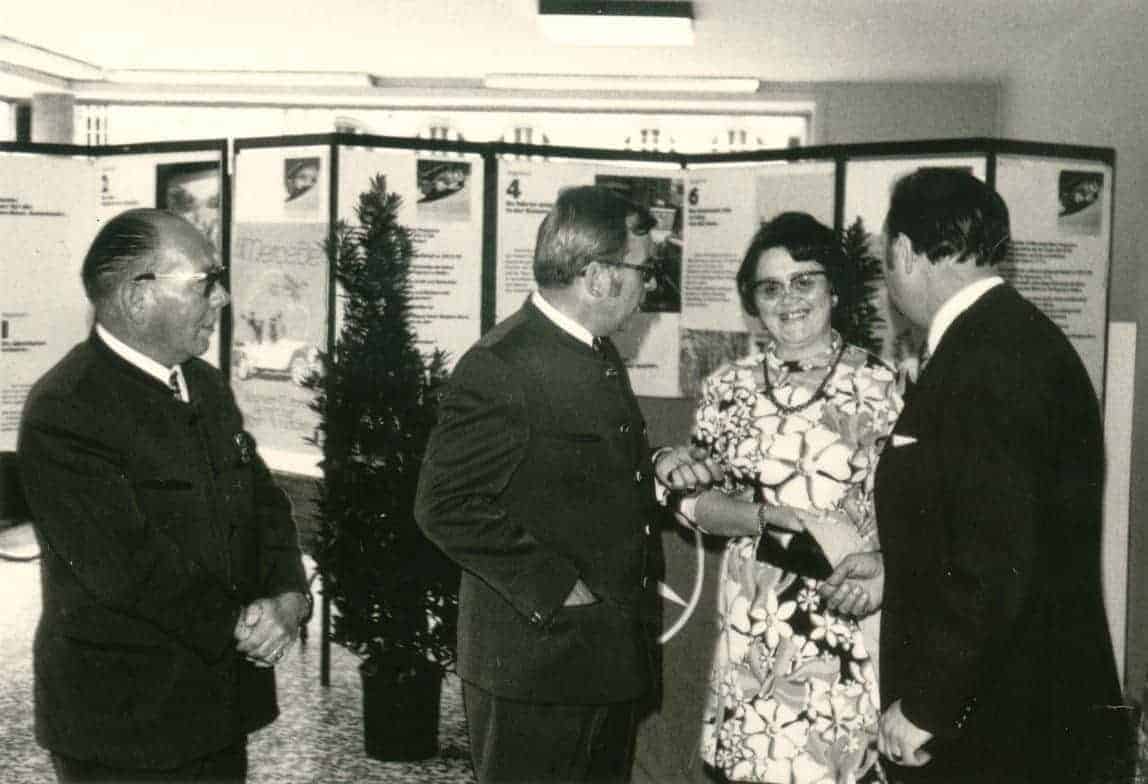 Vier Personen stehen in einem Innenraum und unterhalten sich unter Ihnen Gerda Meurer. Im Hintergrund befinden sich mehrere Stellwände mit Texten und Bildern, vermutlich Teil einer Ausstellung oder Präsentation. Zwischen den Personen und den Stellwänden steht eine Topfpflanze.