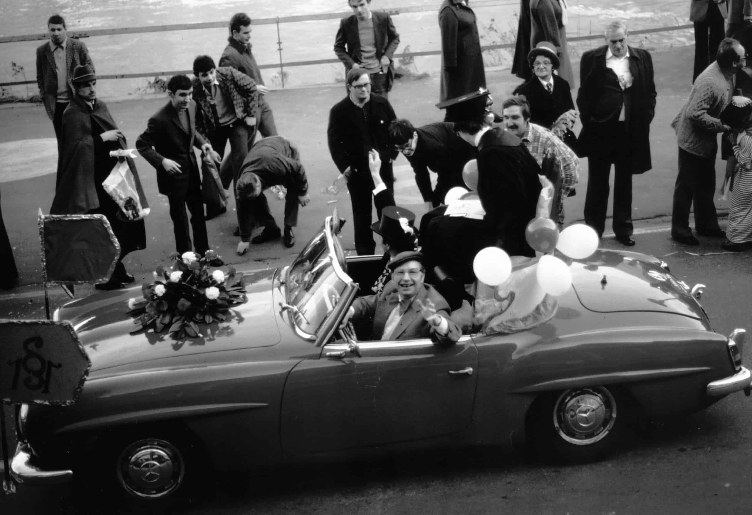 Ein Schwarz-Weiß-Foto zeigt ein Cabriolet, das mit Luftballons und Blumen geschmückt ist. Mehrere Personen stehen um das Fahrzeug herum, teils festlich, teils leger gekleidet. Die Szene wirkt wie eine Feier, möglicherweise eine Hochzeit oder ein Umzug. Im Hintergrund ist ein Geländer zu sehen, dahinter vermutlich Wasser.