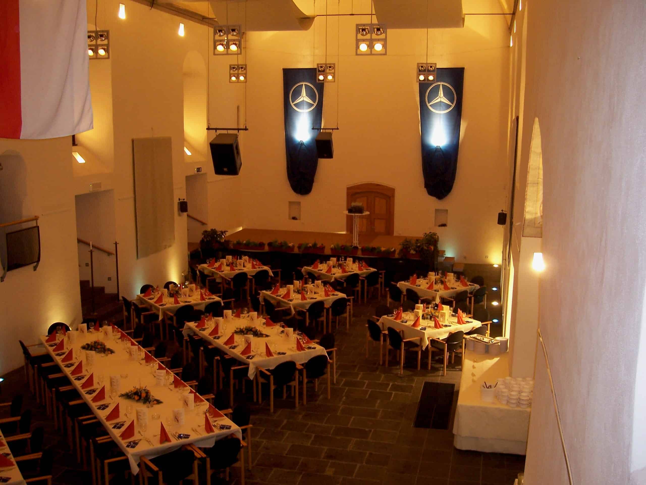 Ein großer, hell erleuchteter Festsaal ist für eine Veranstaltung vorbereitet. Mehrere lange Tische mit weißen Tischdecken und roten Servietten sind in Reihen angeordnet. Die Tische sind mit Tellern, Gläsern und Besteck gedeckt und von schwarzen Stühlen umgeben. Am Ende des Saals befindet sich eine Bühne mit zwei großen blauen Bannern mit Mercedes-Benz-Logo. Die Wände sind weiß, und von der Decke hängen mehrere Leuchten. Links ist teilweise eine Flagge zu sehen.