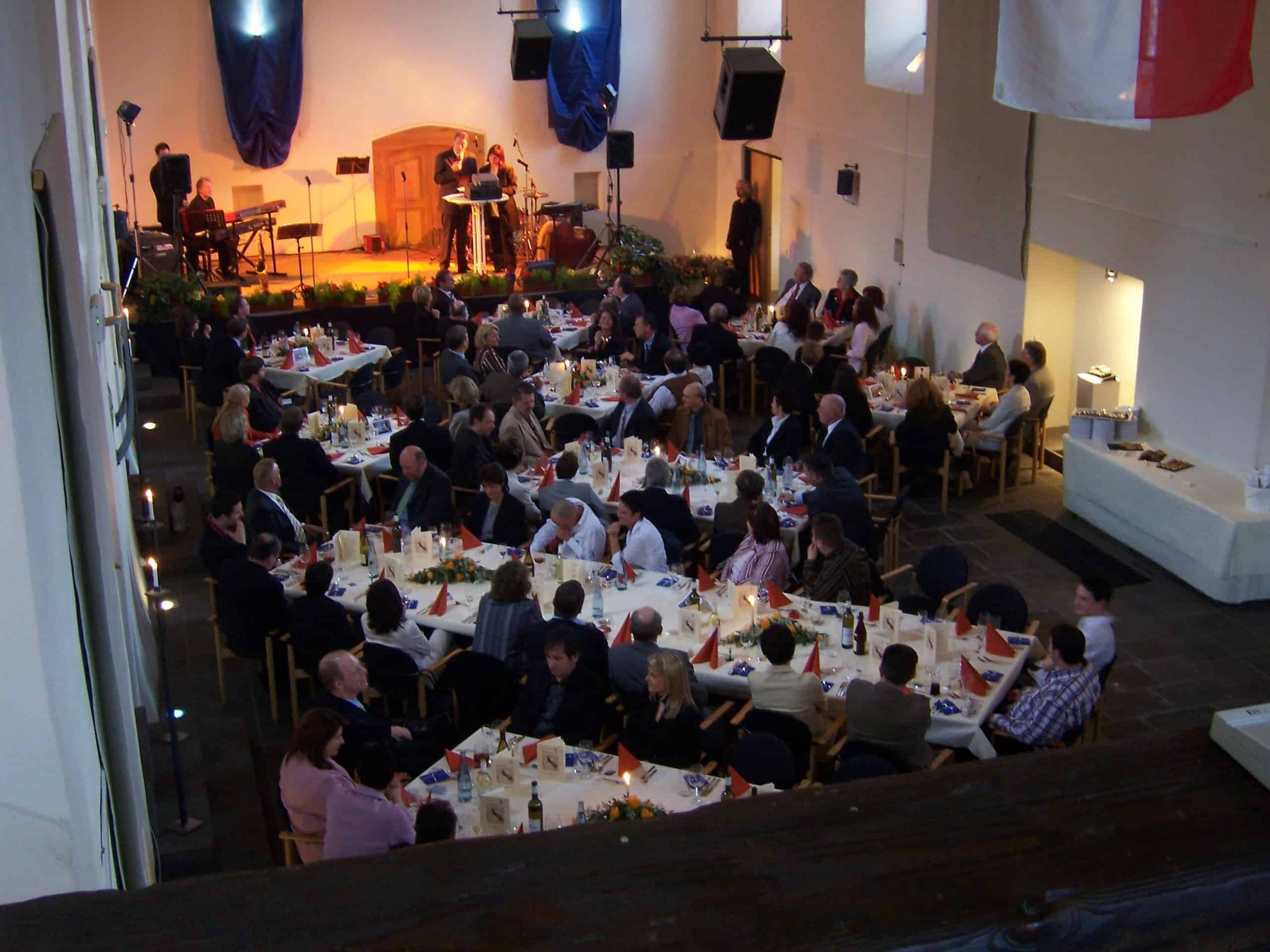 Eine große Veranstaltung mit vielen Personen, die an runden Tischen mit weißen Tischdecken sitzen. Die Tische sind mit Tellern, Gläsern und Flaschen gedeckt. Im Vordergrund befindet sich eine Bühne mit Musikern – eine Person spielt Keyboard, eine andere spricht oder singt ins Mikrofon. Der Saal hat hohe Decken, blaue Wandbehänge und große Lautsprecher, die von der Decke hängen.