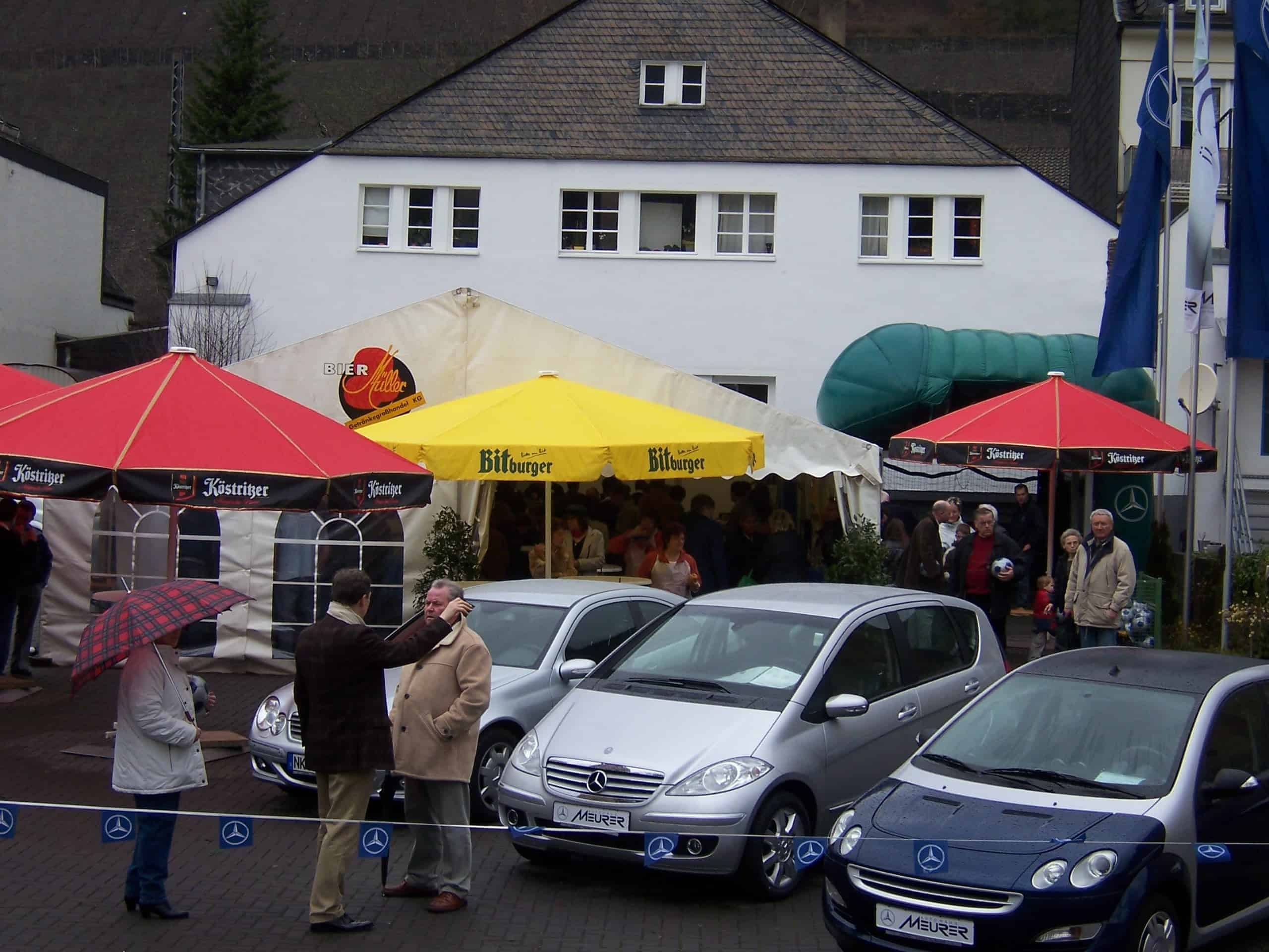 Ein Außenevent mit mehreren Personen, die sich unter großen Sonnenschirmen der Marken „Köstritzer“ und „Bitburger“ versammeln. Unter den Schirmen steht ein Zelt mit der Aufschrift „Bier“. Davor sind zwei silberne Mercedes-Benz Fahrzeuge nebeneinander geparkt. Der Bereich ist mit einem blauen Seil mit Mercedes-Benz-Logo abgesperrt. Im Hintergrund befindet sich ein weißes Gebäude mit mehreren Fenstern und rechts davon Flaggenmasten.