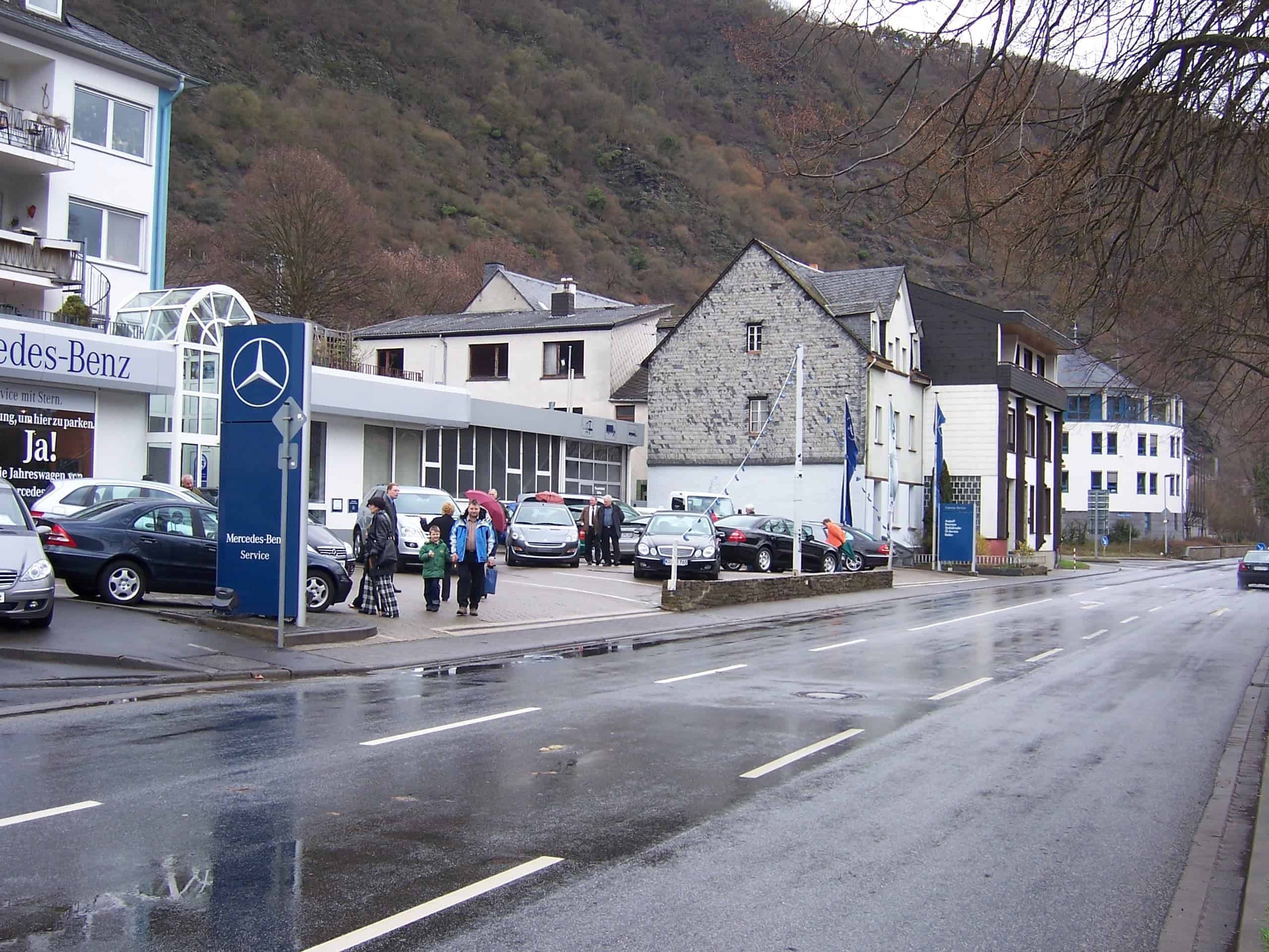 Ein Mercedes-Benz Autohaus an einer Straße mit mehreren geparkten Fahrzeugen und Personen vor dem Gebäude. Im Hintergrund sind Wohnhäuser und ein bewaldeter Hang zu sehen. Die Straße ist nass, was auf kürzlichen Regen hinweist. Das Mercedes-Benz-Logo und entsprechende Beschilderung sind gut sichtbar am Eingang angebracht.