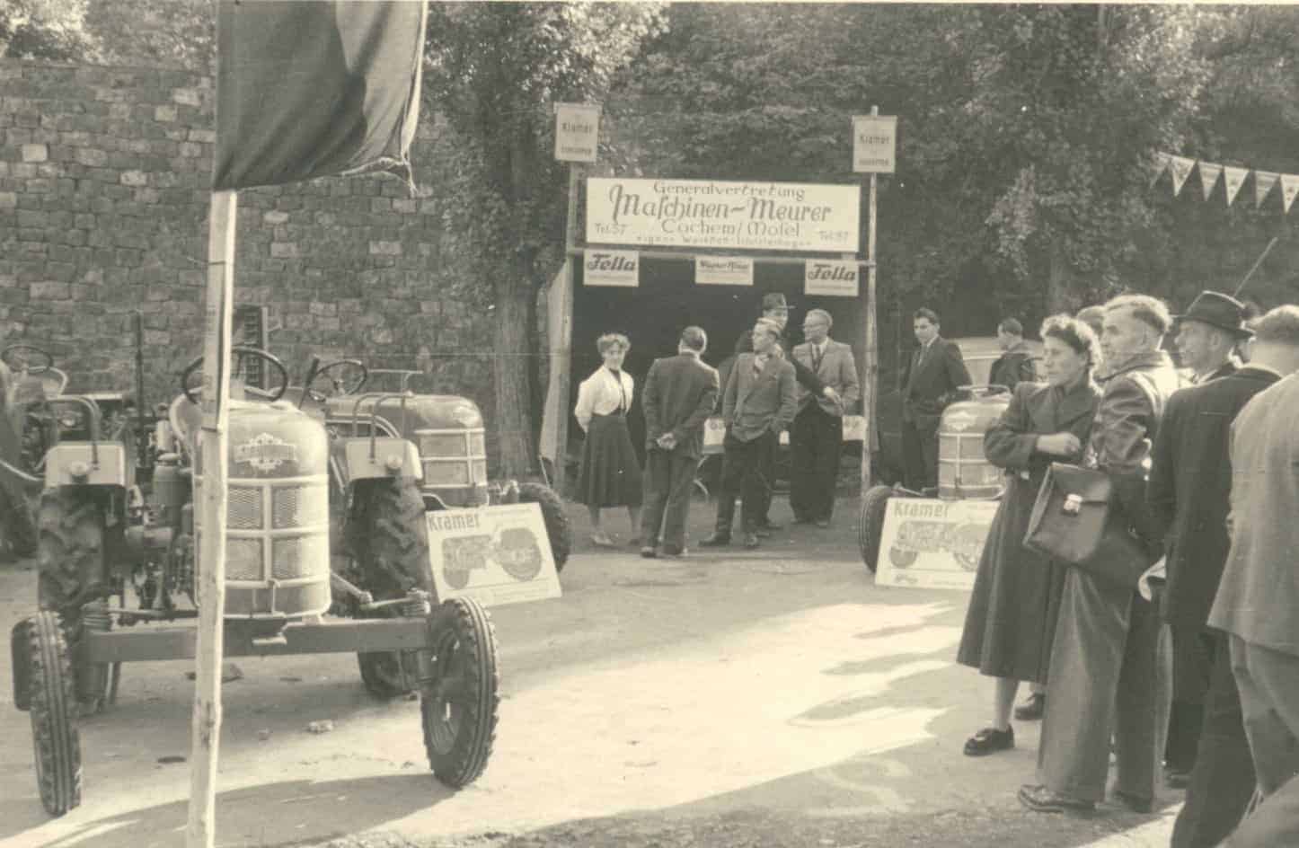 Ein historisches Foto zeigt eine Gruppe von Menschen vor einem Messestand mit der Aufschrift „Generalvertretung Maschinen-Meurer Cochem (Mosel)“. Links und rechts sind Werbeschilder der Marken „Fella“ und „Kramer“ zu sehen. Vor dem Stand stehen zwei Traktoren, während mehrere Personen das Angebot betrachten oder sich unterhalten. Die Szene spielt sich im Freien ab, vermutlich auf einer landwirtschaftlichen Ausstellung.