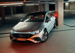 Ein silberner Mercedes-Benz EQS steht mit eingeschaltetem Licht in einer Tiefgarage. Das Kennzeichen lautet „BB EQ 935E“. Zwei Personen sind im Fahrzeuginneren sichtbar. Die Umgebung besteht aus Betonpfeilern und -wänden, beleuchtet durch Deckenlampen. Das Fahrzeug hatte Kontakt zu einem der Betonpfeiler und benötigt das Mercedes-Benz Programm SmallRepair.