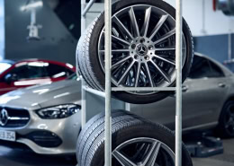 In einer Werkstatt stehen im Hintergrund zwei Mercedes-Benz Fahrzeuge, eines silbern und eines rot. Im Vordergrund befindet sich ein Metallregal mit drei montierten Mercedes-Benz-Leichtmetallrädern. Die Umgebung ist sauber und ordentlich, mit sichtbarem Werkstattequipment.