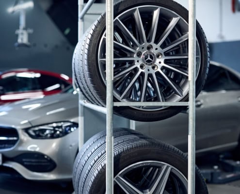 In einer Werkstatt stehen im Hintergrund zwei Mercedes-Benz Fahrzeuge, eines silbern und eines rot. Im Vordergrund befindet sich ein Metallregal mit drei montierten Mercedes-Benz-Leichtmetallrädern. Die Umgebung ist sauber und ordentlich, mit sichtbarem Werkstattequipment.