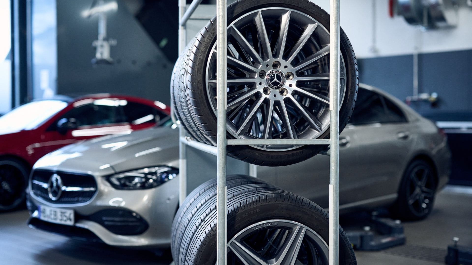 In einer Werkstatt stehen im Hintergrund zwei Mercedes-Benz Fahrzeuge, eines silbern und eines rot. Im Vordergrund befindet sich ein Metallregal mit drei montierten Mercedes-Benz-Leichtmetallrädern. Die Umgebung ist sauber und ordentlich, mit sichtbarem Werkstattequipment.