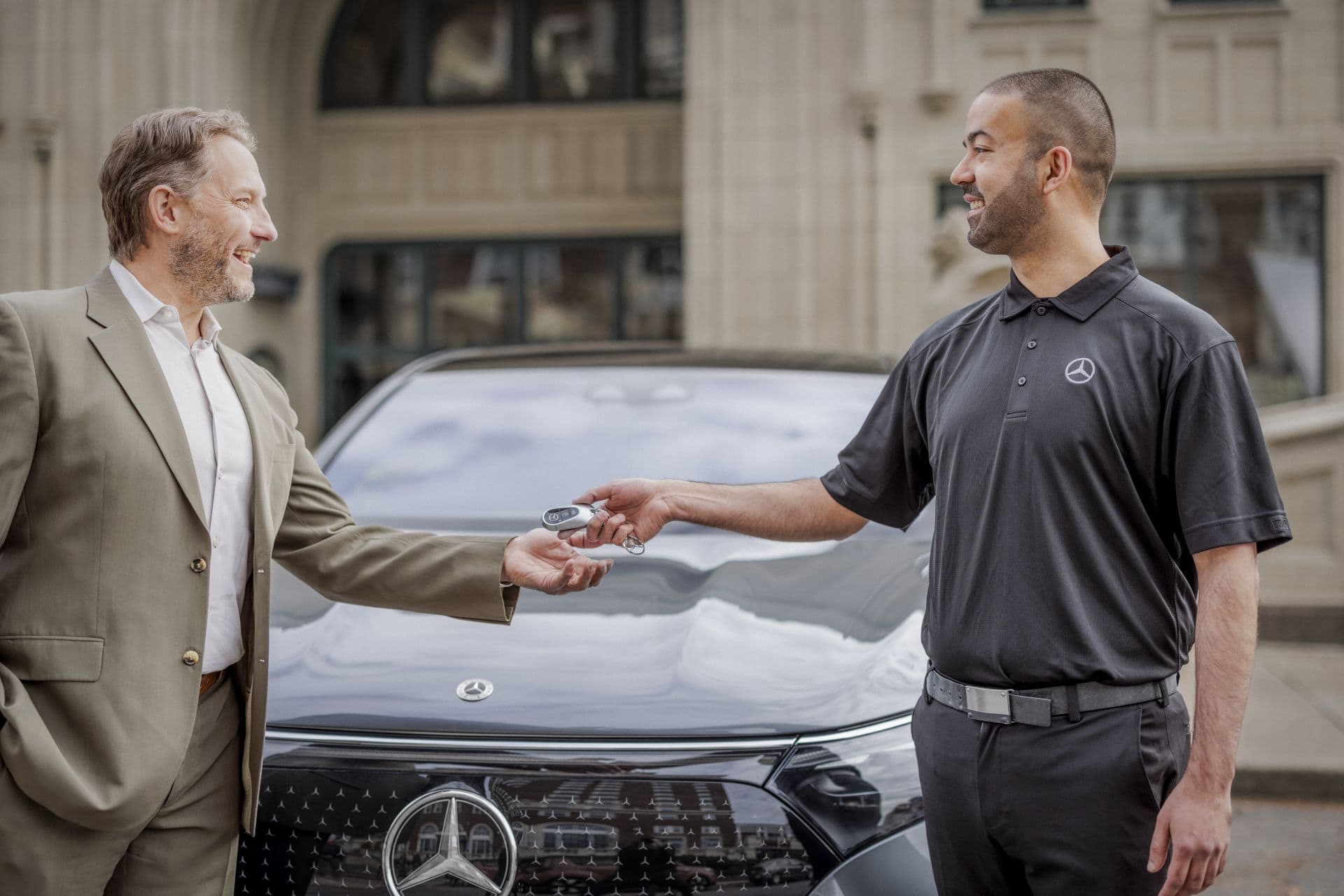 Zwei Personen stehen vor einem Mercedes-Benz Fahrzeug. Eine Person im Anzug übergibt einem anderen, der ein schwarzes Polohemd mit Mercedes-Benz-Logo trägt, einen Autoschlüssel. Im Hintergrund befindet sich ein Gebäude mit großen Fenstern.