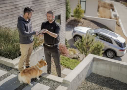 Zwei Personen stehen auf einer Außentreppe. Eine Person übergibt der anderen einen Autoschlüssel. Neben ihnen steht ein Corgi-Hund. Im Hintergrund ist ein SUV vor einem Wohnhaus geparkt.