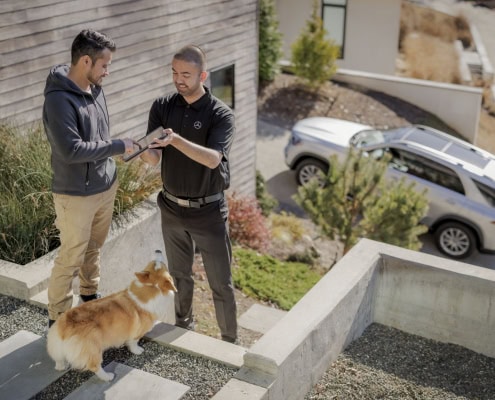 Zwei Personen stehen auf einer Außentreppe. Eine Person übergibt der anderen einen Autoschlüssel. Neben ihnen steht ein Corgi-Hund. Im Hintergrund ist ein SUV vor einem Wohnhaus geparkt.