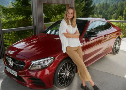 Eine Person lehnt an einem roten Mercedes-Benz. Das Fahrzeug trägt das Kennzeichen „ABR 5119“. Im Hintergrund sind Bäume und Grünflächen zu sehen, was auf eine Außenaufnahme hinweist.