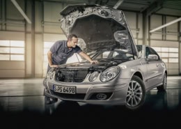 Eine Person inspiziert den Motorraum eines silbernen Mercedes-Benz mit geöffneter Motorhaube. Das Fahrzeug steht in einer gut beleuchteten Werkstatt oder Garage. Das Kennzeichen lautet „S PS 6498“.