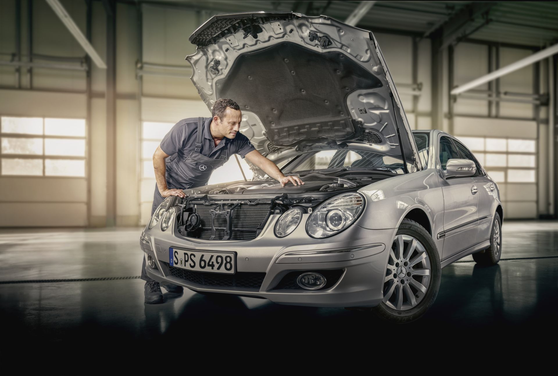 Eine Person inspiziert den Motorraum eines silbernen Mercedes-Benz mit geöffneter Motorhaube. Das Fahrzeug steht in einer gut beleuchteten Werkstatt oder Garage. Das Kennzeichen lautet „S PS 6498“.
