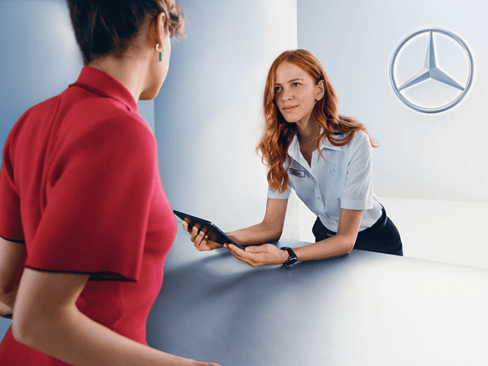 Zwei Personen stehen an einem Tresen. Eine Person in roter Kleidung steht mit dem Rücken zur Kamera und blickt auf den Tresen. Die andere Person, mit langen Haaren und weißem Hemd samt Namensschild, steht hinter dem Tresen und hält ein Tablet in der Hand. Im Hintergrund ist das Mercedes-Benz-Logo an der Wand sichtbar.