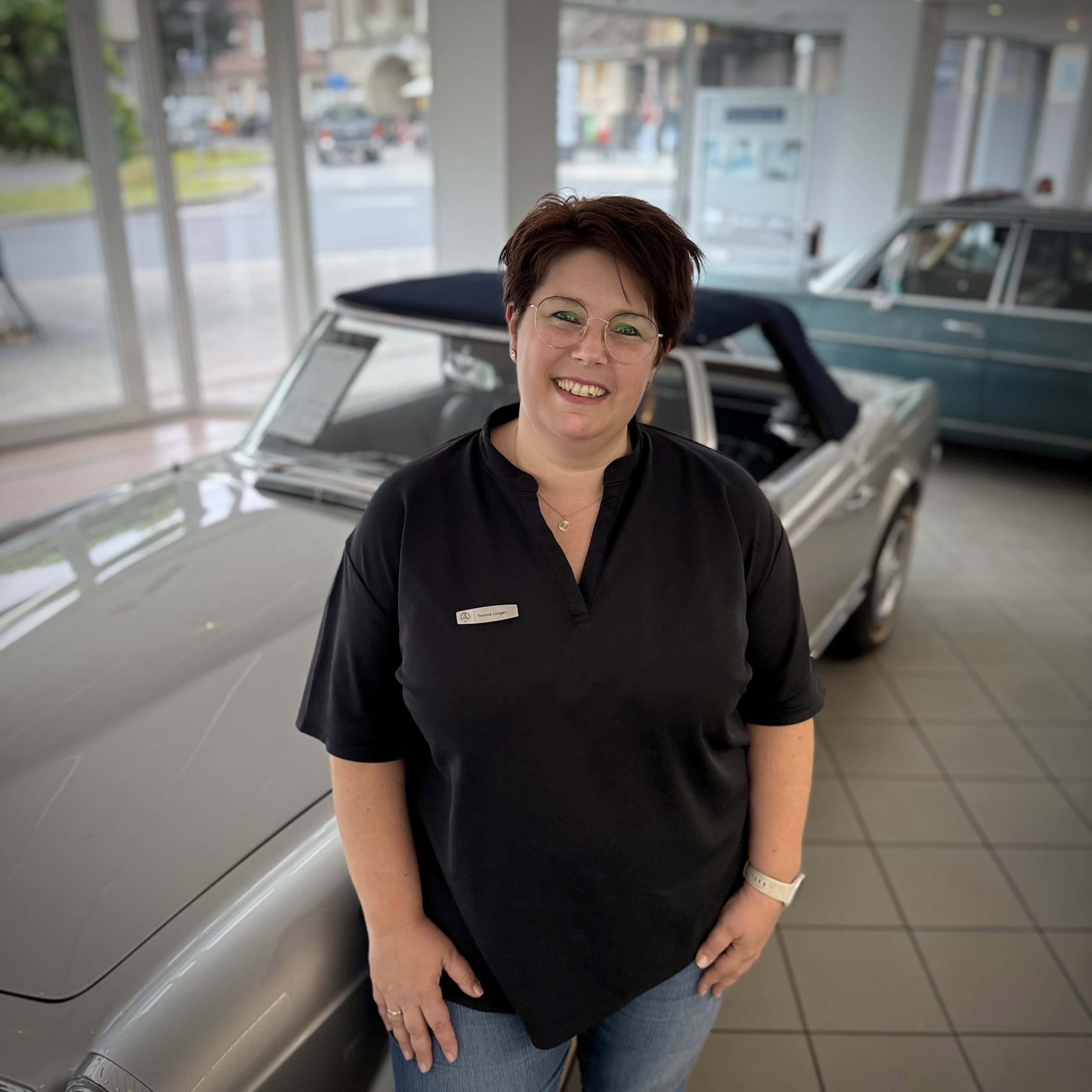 Eine Person mit kurzem Haar und schwarzem Shirt mit Namensschild steht vor einem klassischen Cabrio in einem Ausstellungsraum. Im Hintergrund ist ein weiteres klassisches Fahrzeug zu sehen. Der Raum hat große Fenster mit Blick auf die Straße draußen.