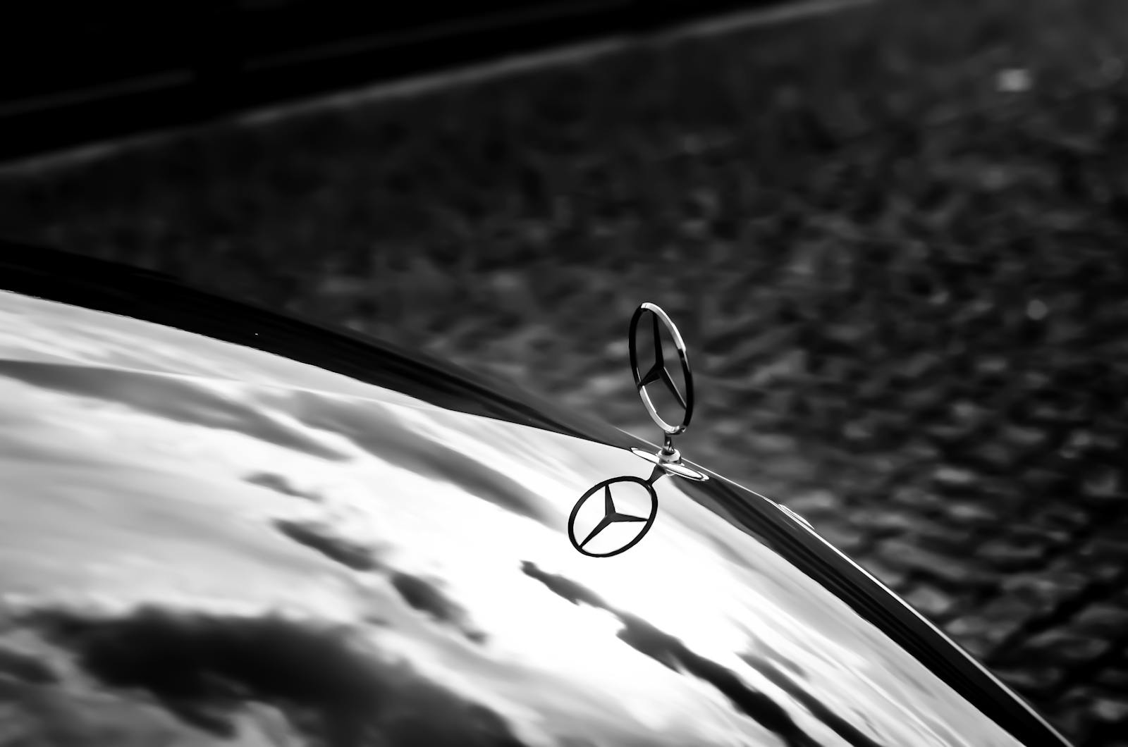 Mercedes-Benz-Kühlerfigur in Form eines dreizackigen Sterns im Kreis, montiert auf der Motorhaube eines Fahrzeugs. Im Hintergrund ist ein Kopfsteinpflaster zu sehen, auf der glänzenden Oberfläche der Motorhaube spiegeln sich Wolken.